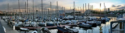 Panoramafoto eines dicht belegten Yachthafens mit zahlreichen Segelbooten und Motorbooten vor Sonnenuntergang, im Hintergrund Gebäude und ein Hügel.