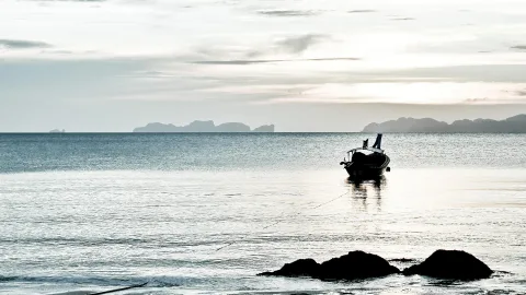 Ein kleines Boot liegt ruhig auf dem spiegelnden Meer, im Hintergrund zeichnen sich dunkle Inseln am Horizont ab, im Vordergrund ragen Felsen aus dem Wasser.