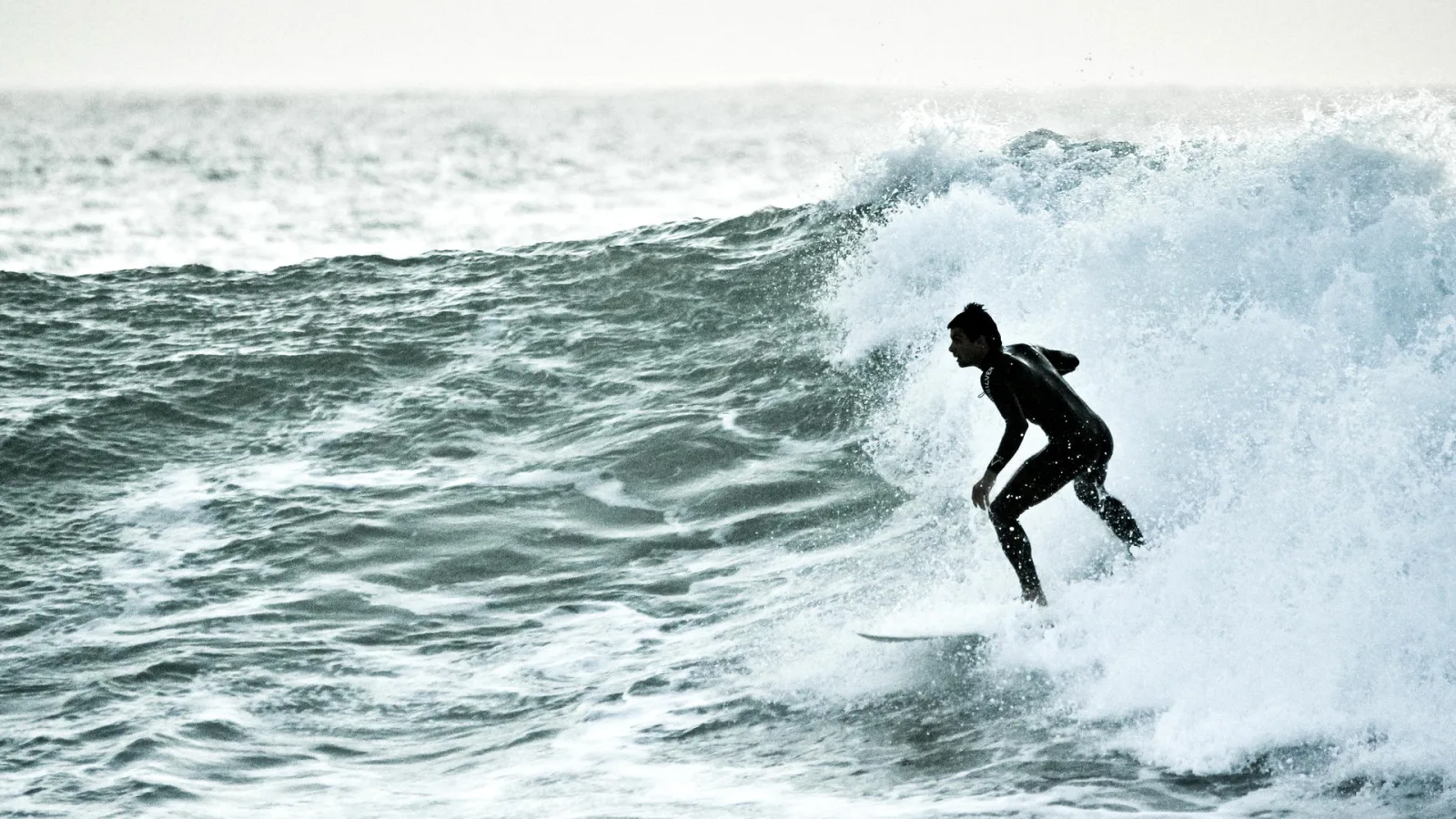 Ein Surfer im schwarzen Neoprenanzug reitet eine Welle, weiße Gischt spritzt um ihn herum.