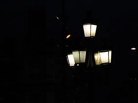 Eine mehrarmige Straßenlaterne mit vier leuchtenden Laternen vor einem dunklen Nachthimmel, links im Schatten die Silhouette eines Gebäudes.
