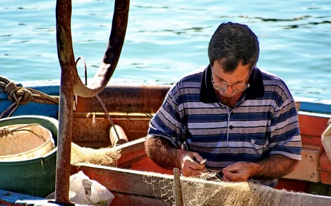 Ein Fischer mit Brille und gestreiftem Poloshirt flickt auf einem Holzboot ein Fischernetz, im Hintergrund glitzert das türkisfarbene Meer.