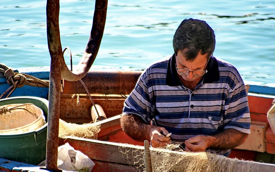Ein Fischer mit Brille und gestreiftem Poloshirt flickt auf einem Holzboot ein Fischernetz, im Hintergrund glitzert das türkisfarbene Meer.