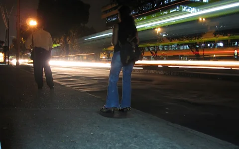 Nächtliche Straße mit langen Lichtspuren vorbeifahrender Fahrzeuge. Vorne steht eine Frau mit Rucksack und Jeans am Bordstein, links daneben ein Mann in heller Hemd. Im Hintergrund ein beleuchtetes Gebäude mit grünen Akzenten.