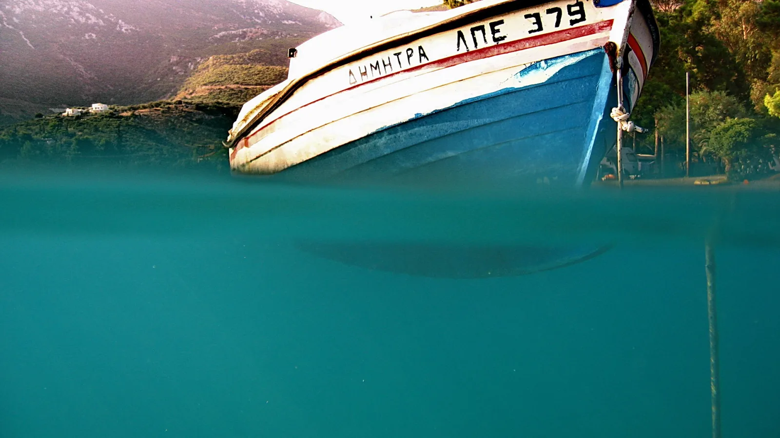 Ein blau-weiß lackiertes Fischerboot mit griechischer Beschriftung ragt an der Wasseroberfläche aus türkisfarbenem Meer, im Hintergrund bewaldete Hügel und ein weißes Haus.