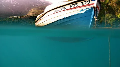 Ein blau-weiß lackiertes Fischerboot mit griechischer Beschriftung ragt an der Wasseroberfläche aus türkisfarbenem Meer, im Hintergrund bewaldete Hügel und ein weißes Haus.