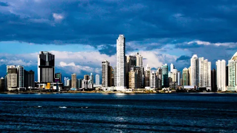 Skyline einer Großstadt mit dicht stehenden Hochhäusern am Wasser unter dunkelblauem Wolkenhimmel, im Vordergrund die dunkle Bucht.