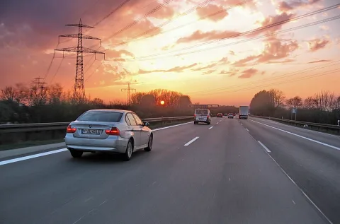 Silberner BMW und weiße Transporter auf einer Autobahn bei Sonnenuntergang, daneben Strommasten und Bäume.