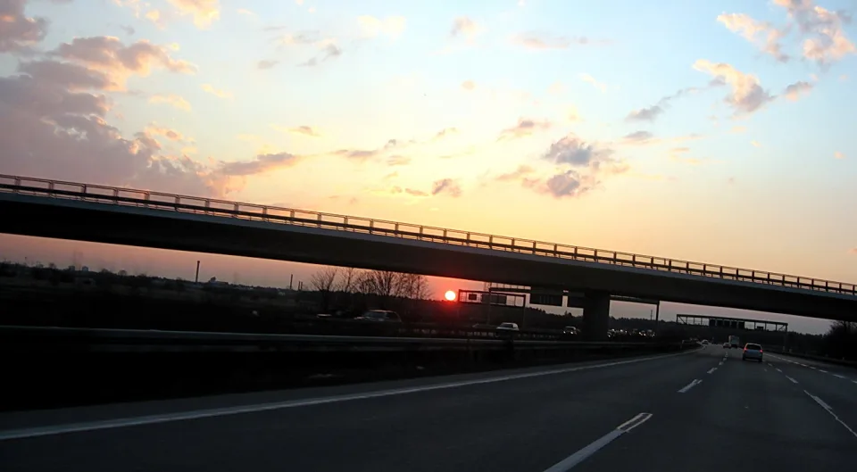 Autobahn bei Sonnenuntergang, eine Brücke überspannt die Fahrbahn, am Horizont steht die rote Sonne tief über dem Land.