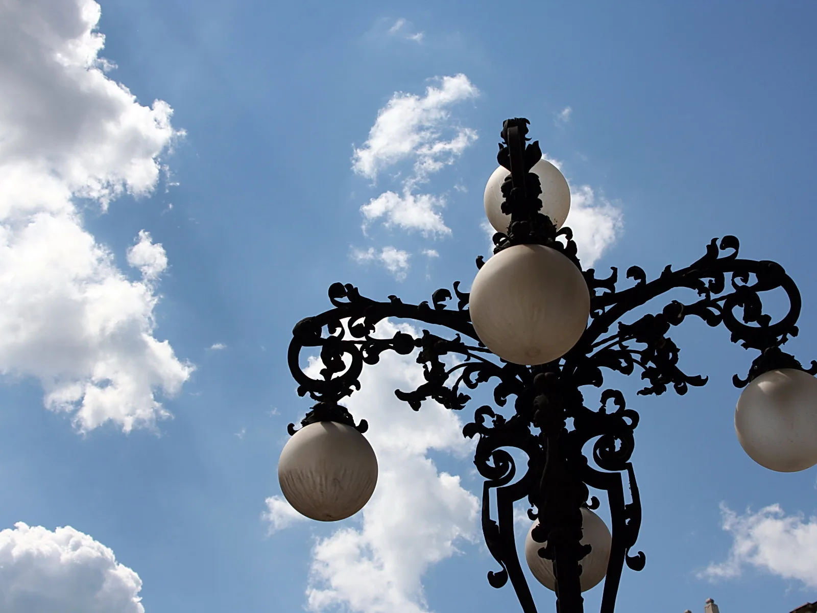 Schwarze schmiedeeiserne Straßenlaterne mit vier milchweißen Glaskugeln vor blauem Himmel mit Wolken, von unten fotografiert.