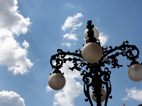 Schwarze schmiedeeiserne Straßenlaterne mit vier milchweißen Glaskugeln vor blauem Himmel mit Wolken, von unten fotografiert.