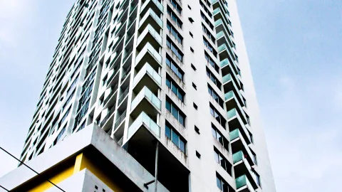 Weißes Hochhaus mit vielen Balkonen, die mit mintgrünen Glasbrüstungen versehen sind, aufgenommen von unten vor hellblauem Himmel. Im unteren linken Bereich ein gelb akzentuierter Betonvorsprung.