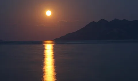 Ein heller Mond steht in der Dämmerung über einer weiten, glatten Wasserfläche. Auf dem Wasser zieht sich eine goldene Spiegelung senkrecht nach unten, rechts zeichnet sich eine dunkle Gebirgskette am Horizont ab.