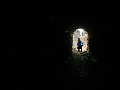 Eine Person in blauer Jacke und Mütze hockt im Ausgang eines dunklen, gewölbten Tunnels, dahinter sind helle felsige Wände zu sehen.