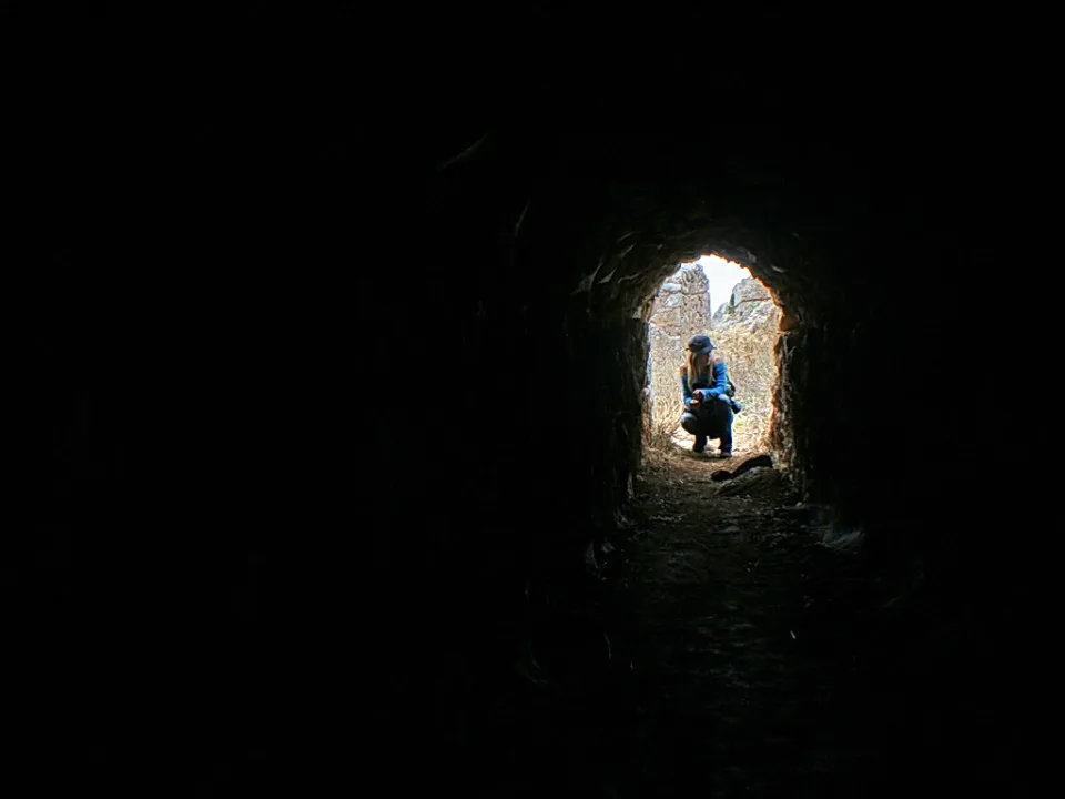 Eine Person in blauer Jacke und Mütze hockt im Ausgang eines dunklen, gewölbten Tunnels, dahinter sind helle felsige Wände zu sehen.