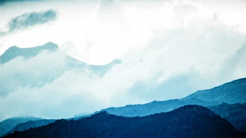 Mehrere blaugraue Gebirgskämme schieben sich von unten ins Bild, darüber hängen dichte weiße Wolken und Nebel, hinter denen ein heller Himmel durchscheint.