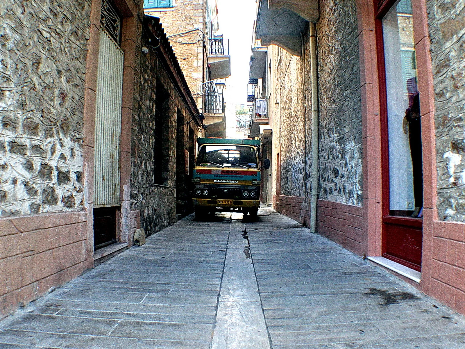 Ein grün-gelber Lkw steht am Ende einer schmalen Gasse zwischen Steinmauern und rot gestrichenen Hauswänden, das Pflaster führt geradeaus zu ihm.