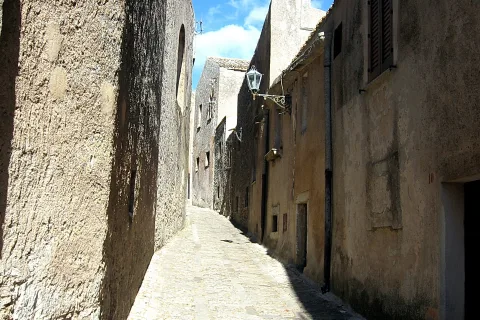 Schmale, kopfsteingepflasterte Gasse zwischen sandfarbenen Mauern unter blauem Himmel, an der linken Wand hängt eine Laterne.