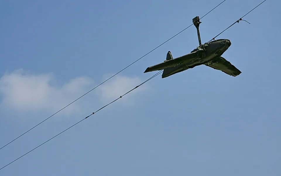 Ein grau-grünes Modellflugzeug hängt kopfüber an einer Stromleitung vor blauem Himmel mit einzelner Wolke links.
