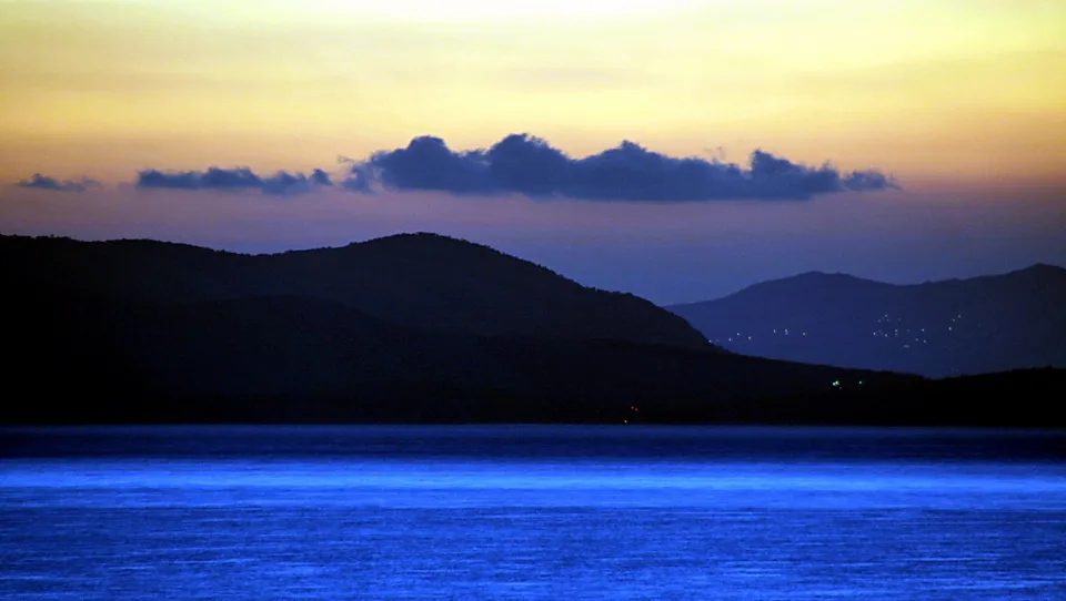 Dunkle Berge zeichnen sich vor einem gelb-orangen Abendhimmel ab, darunter leuchtet die Wasserfläche blau.