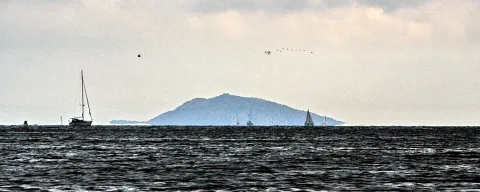 Dunkles, bewegtes Meer im Vordergrund, dahinter mehrere Segelboote und eine bewaldete Insel unter grauem, bewölktem Himmel.