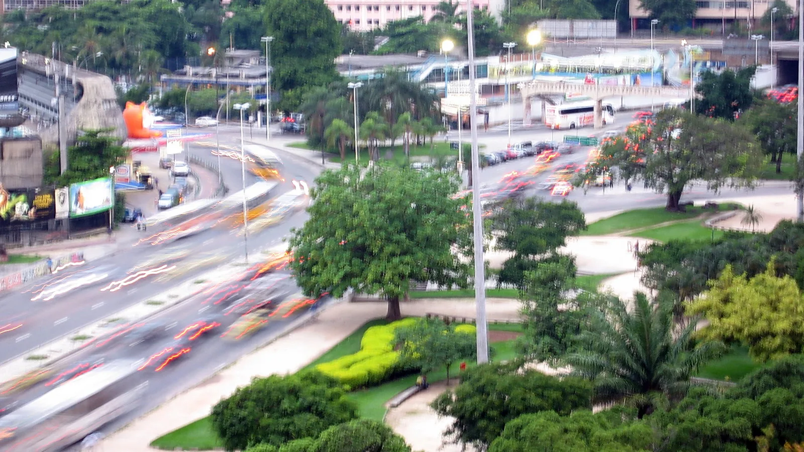 Langzeitbelichtung einer mehrspurigen Straße mit Lichtspuren von Autos und Bussen, dahinter Bäume und Palmen in Rio de Janeiro.