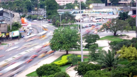 Langzeitbelichtung einer mehrspurigen Straße mit Lichtspuren von Autos und Bussen, dahinter Bäume und Palmen in Rio de Janeiro.