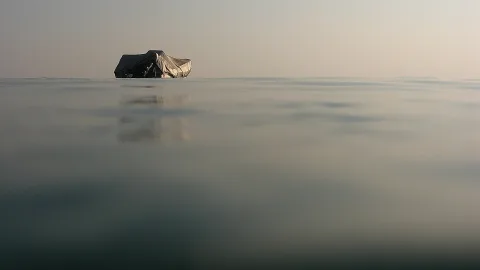 Ein grau abgedecktes Boot schwimmt weit draußen auf einer glatten Wasserfläche unter einem hellen, dunstigen Himmel, das Wasser spiegelt das Boot.