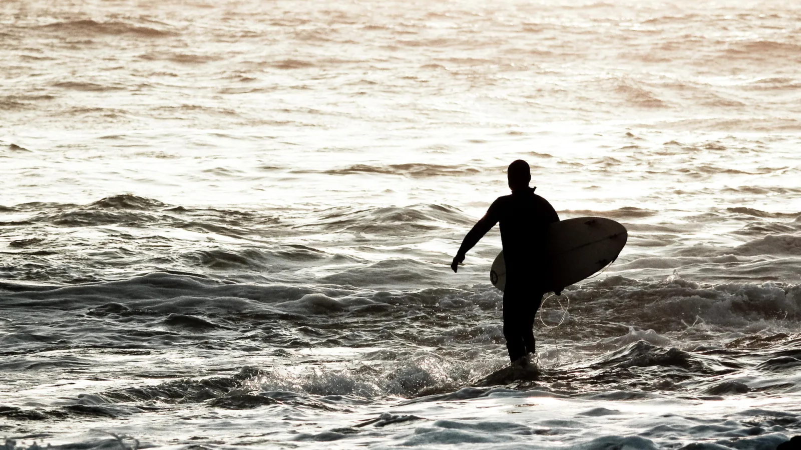 Silhouette eines Surfers mit Brett unterm Arm, der bei Sonnenuntergang durch die Brandung zum Strand watet.