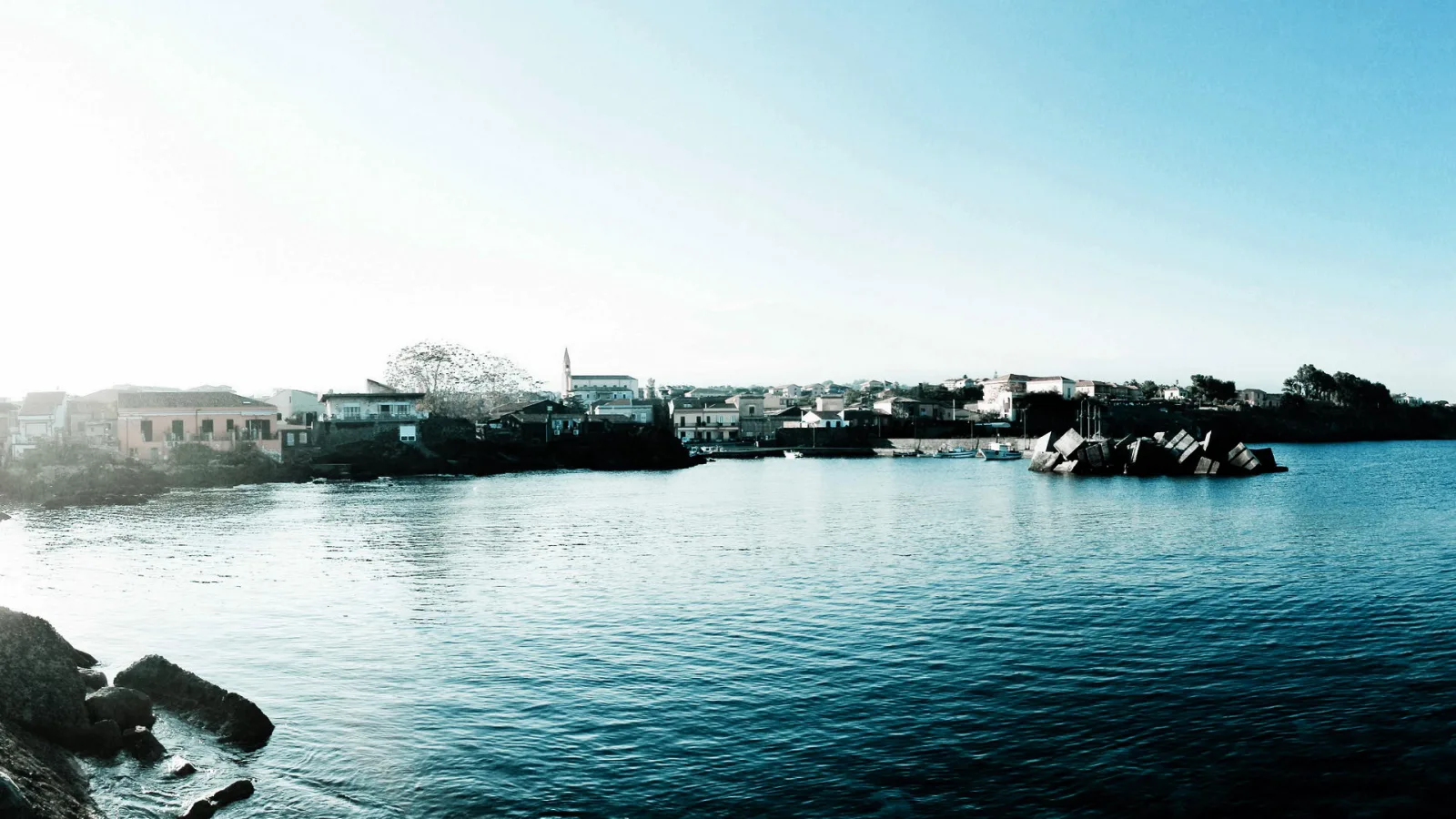 Panoramablick auf eine Hafenbucht mit blauem Wasser, einer Küstenstadt mit weißen und ockerfarbenen Häusern, einem Kirchturm und dunklen Felsblöcken am Wasser unter hellem, diesigem Himmel.