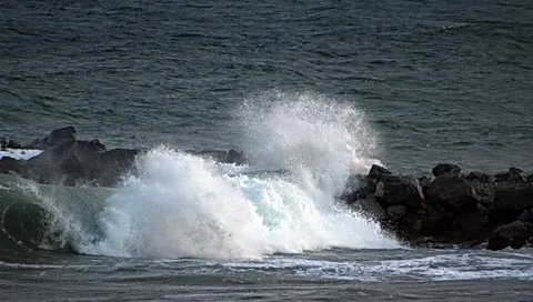 Eine große weiße Welle bricht mit viel Gischt gegen eine Reihe dunkler Felsen am Wellenbrecher, das Meer dahinter ist grau-grün.