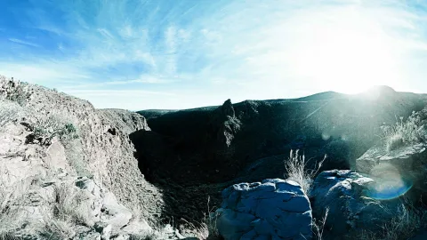 Felsige Schlucht mit Kakteen und trockenen Gräsern im Vordergrund, dahinter ein steiler Felsgrat und Bergrücken unter hellem blauem Himmel mit Sonne rechts oben.