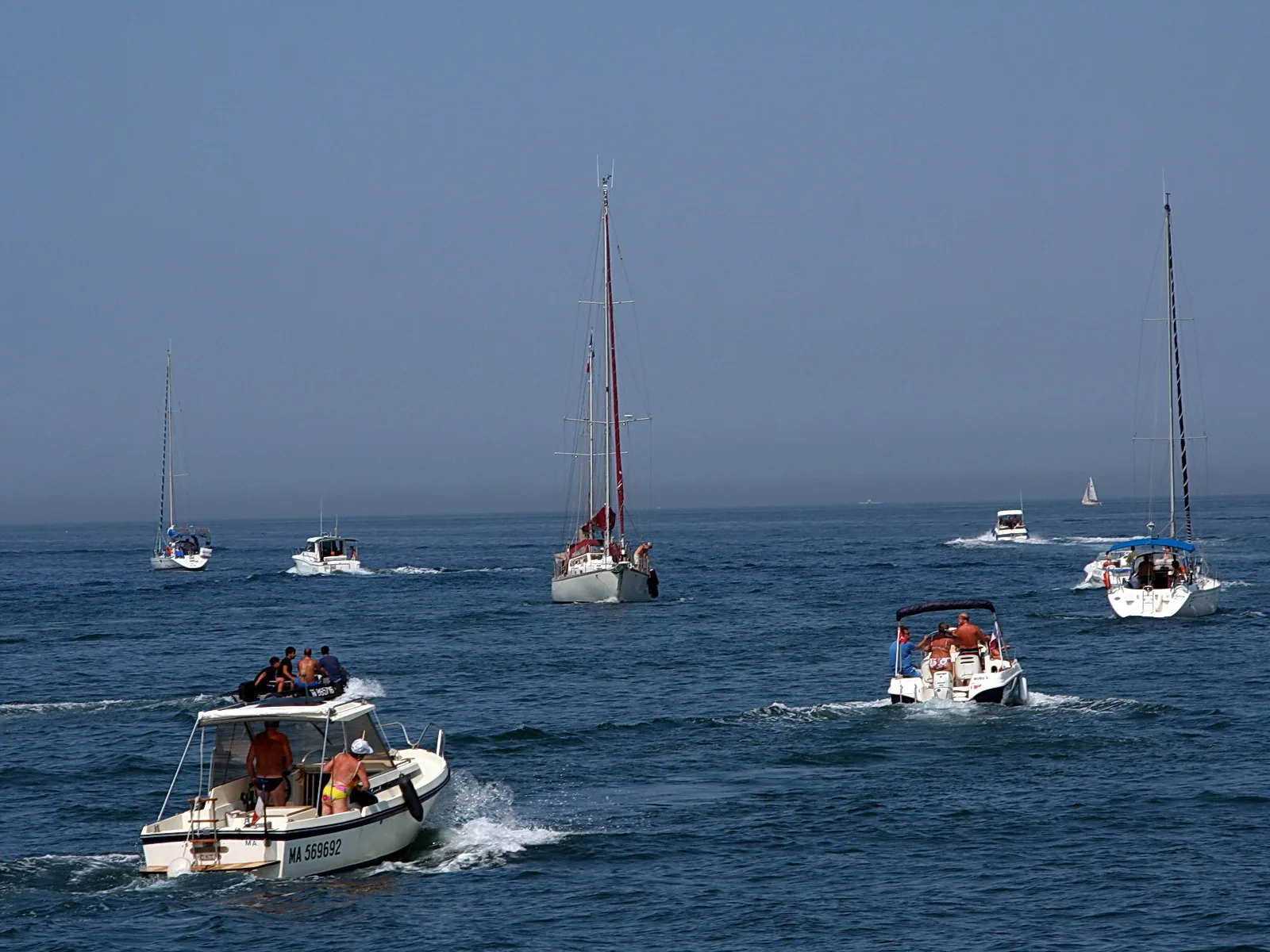 Mehrere Segel- und Motorboote fahren im Abstand über ein blaues Meer, im Vordergrund ein weißes Motorboot mit mehreren Menschen an Bord, im Hintergrund ein Segelboot mit rotem Mast unter dunstigem Himmel.
