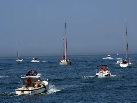 Mehrere Segel- und Motorboote fahren im Abstand über ein blaues Meer, im Vordergrund ein weißes Motorboot mit mehreren Menschen an Bord, im Hintergrund ein Segelboot mit rotem Mast unter dunstigem Himmel.