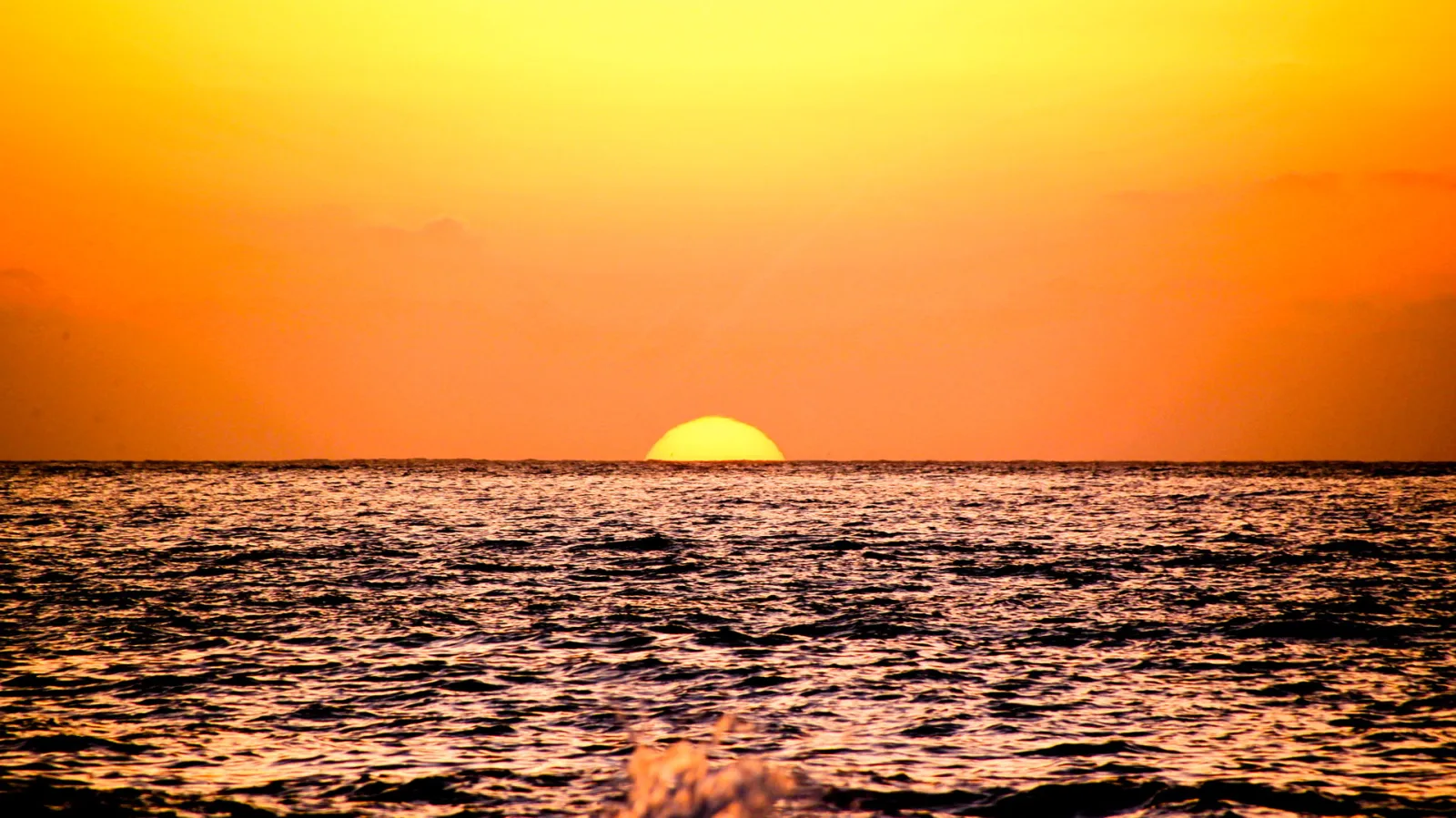 Halb versunkene gelbe Sonne über einem Meereshorizont, darunter dunkle Wellen mit Glitzerreflexen in einem orangefarbenen Himmel