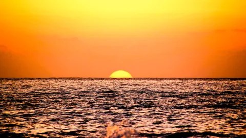 Halb versunkene gelbe Sonne über einem Meereshorizont, darunter dunkle Wellen mit Glitzerreflexen in einem orangefarbenen Himmel