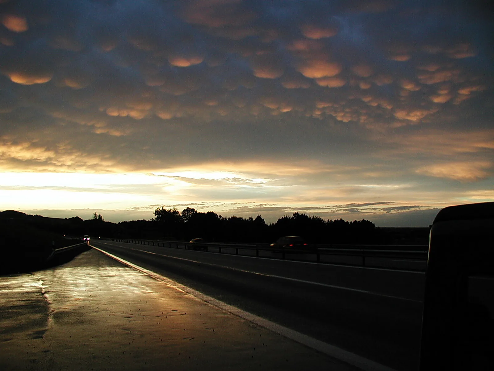 Autobahn bei Sonnenuntergang unter einem Himmel mit wellenförmigen Wolkenbäuchen in Orange und Grau, rechts ein Lkw, im Hintergrund dunkle Bäume.