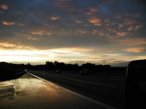 Autobahn bei Sonnenuntergang unter einem Himmel mit wellenförmigen Wolkenbäuchen in Orange und Grau, rechts ein Lkw, im Hintergrund dunkle Bäume.