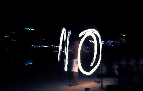 Eine Person steht nachts am Strand und hält eine leuchtende Lichtspur, die einen Kreis und einen senkrechten Strich formt.