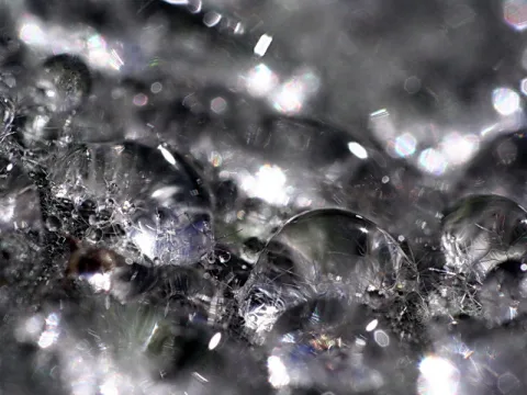 Makroaufnahme mehrerer runder Wassertropfen auf einer grauen, glitzernden Oberfläche mit hellen Lichtreflexen