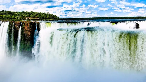 Breiter Wasserfall mit weißer Gischt und Nebel, links ein bewaldeter Felsvorsprung mit kleineren Fällen, darüber blauer Himmel mit Wolken.