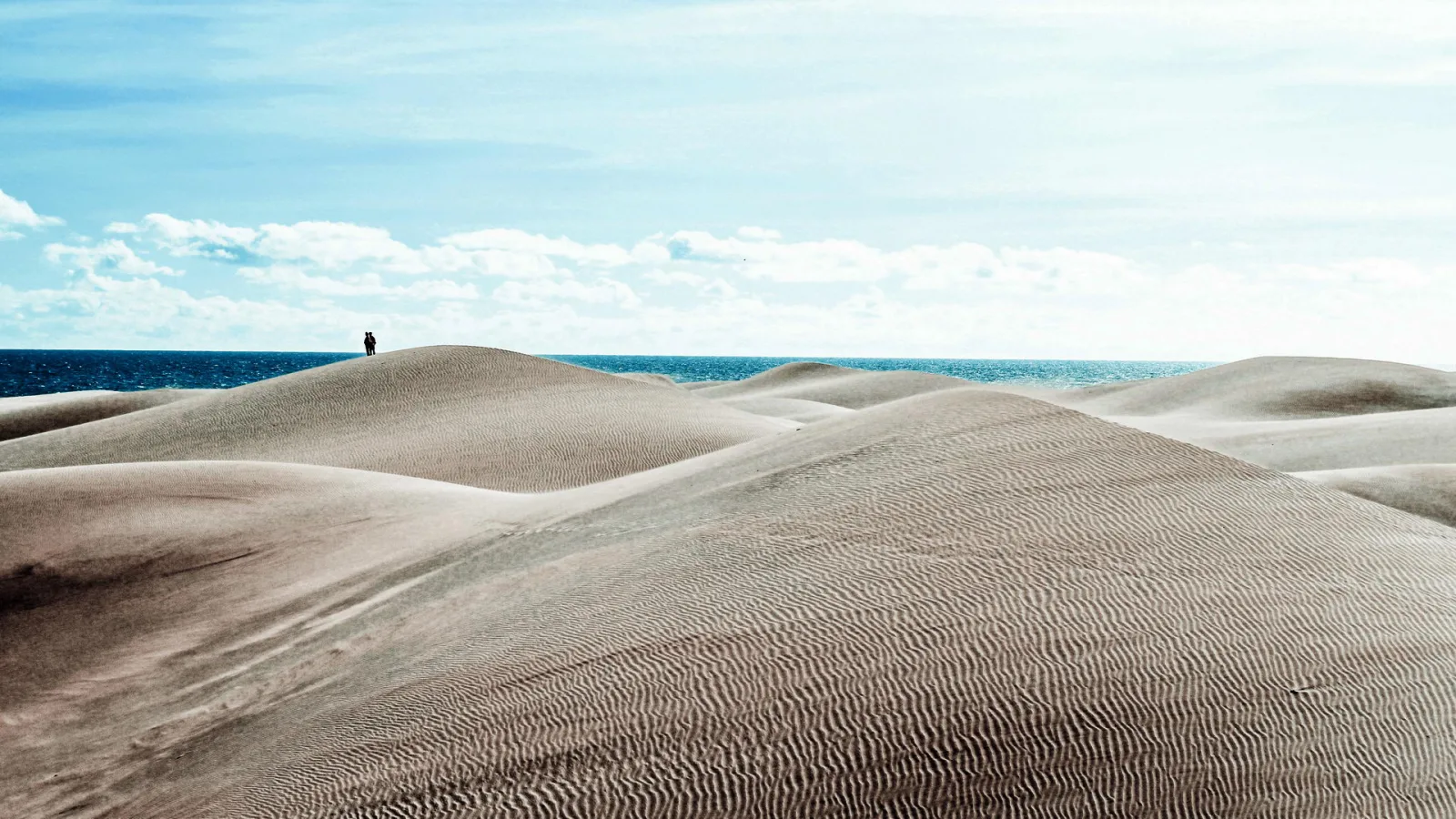 Sanddünen mit welligen Rippelmustern vor blauem Meer unter hellem Himmel, auf einem Dünenkamm stehen zwei kleine Personen.