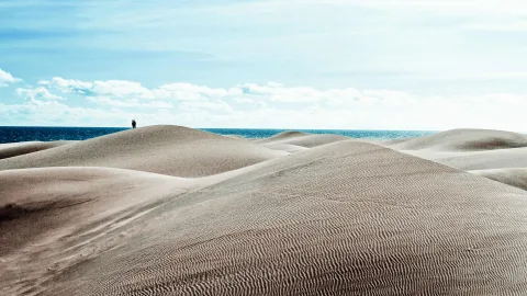 Sanddünen mit welligen Rippelmustern vor blauem Meer unter hellem Himmel, auf einem Dünenkamm stehen zwei kleine Personen.