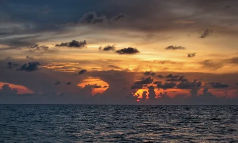 Sonnenuntergang über ruhiger See, Wolken am Abendhimmel leuchten orange und dunkelblau, am Horizont glüht die Sonne zwischen den Wolkenbänken.