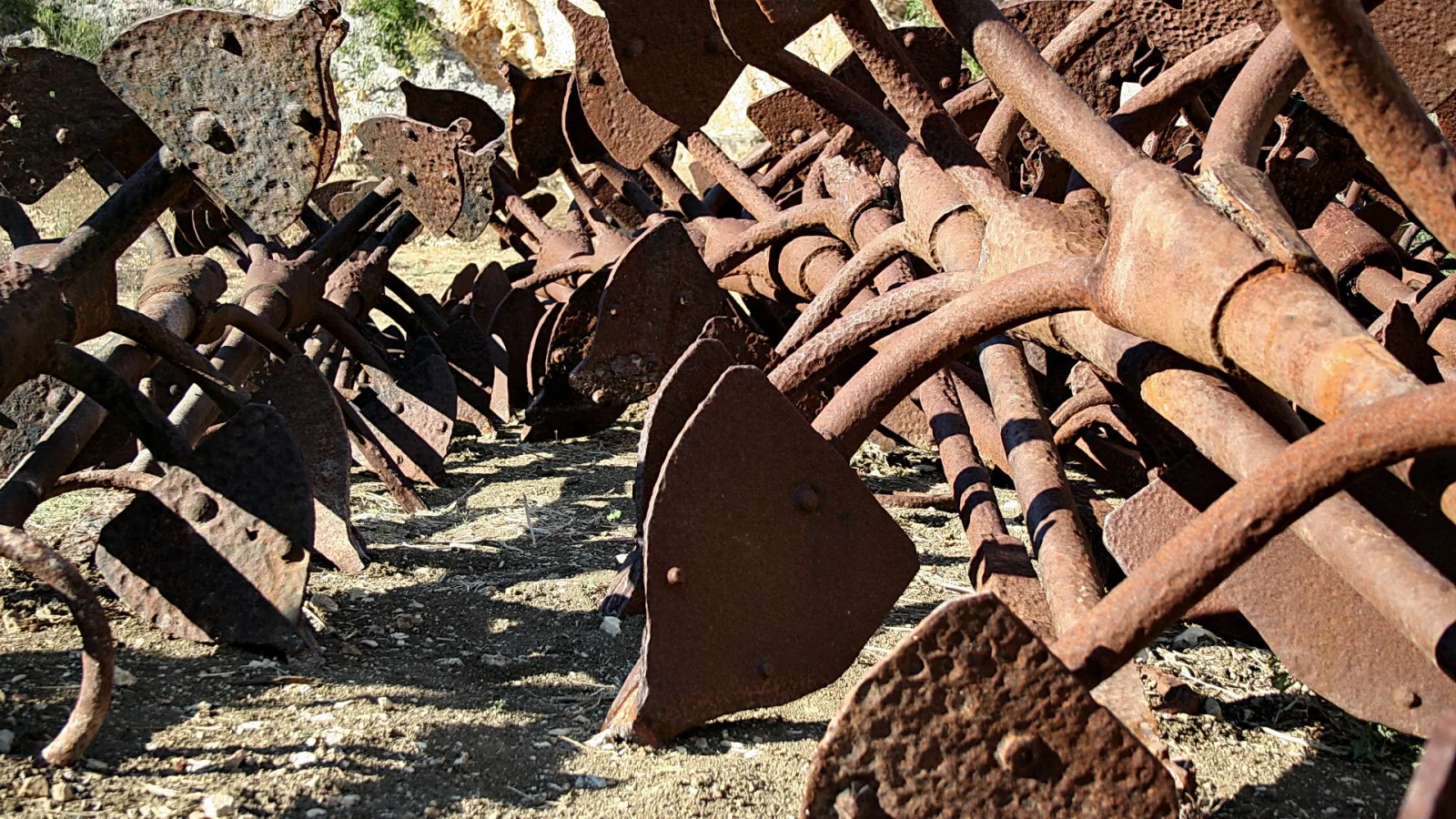 Dicht gestapelte, stark verrostete landwirtschaftliche Geräte mit Metallscheiben und Stangen auf trockenem, staubigem Boden im Sonnenlicht