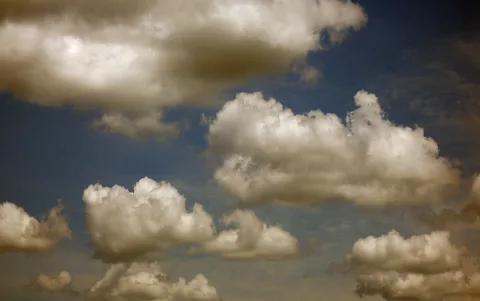 Mehrere weiße Kumuluswolken treiben vor einem tiefblauen Himmel, teils von warmem Licht beschienen.
