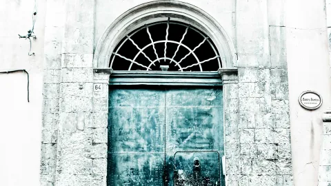 Großes, altes Holztor mit Metallbeschlägen unter einem halbrunden Fenster mit fächerförmigen Sprossen, in einer hellen Steinfassade mit der Hausnummer 64 und einem kleinen Schild Centro Storico.