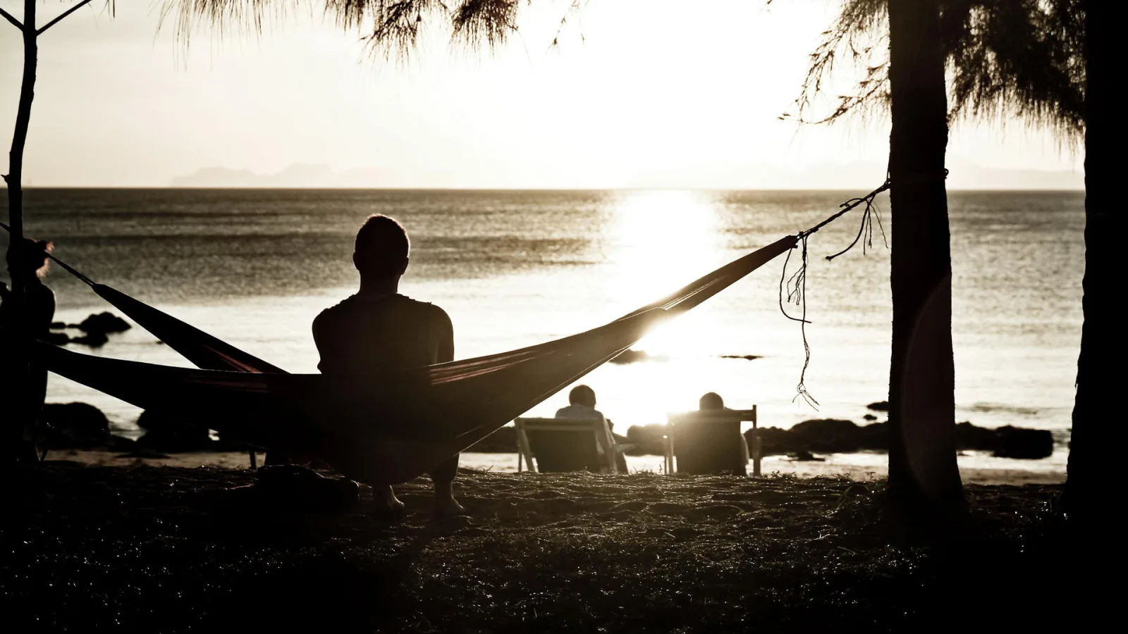 Silhouette eines Menschen in einer Hängematte am Strand, im Hintergrund zwei Personen auf Liegestühlen vor dem Meer bei Sonnenuntergang.