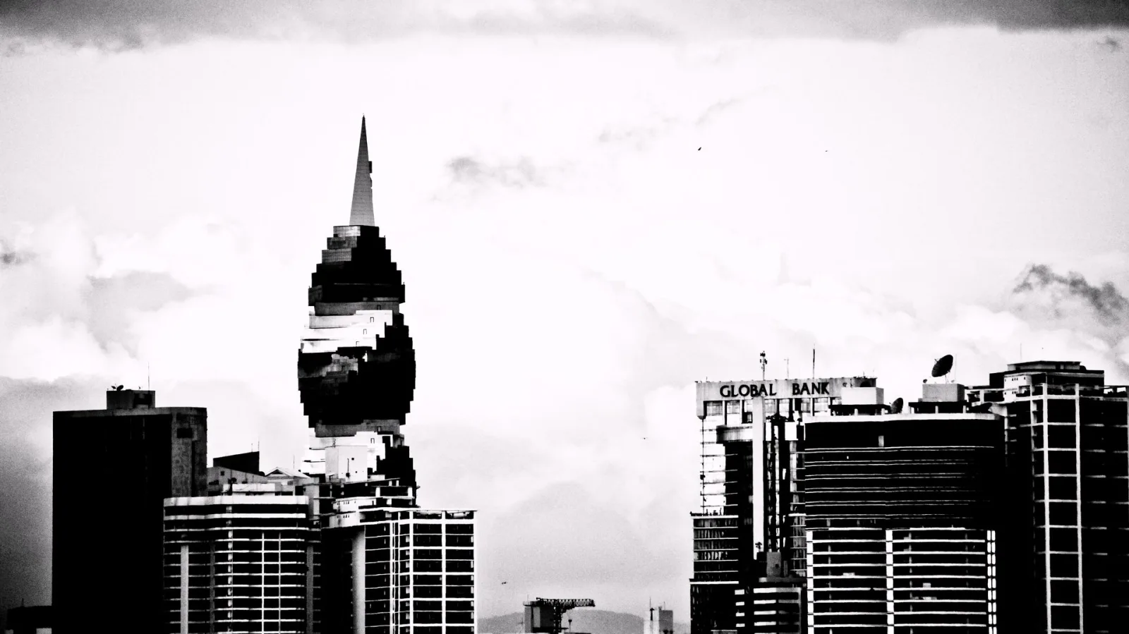 Schwarzweiß-Aufnahme einer Stadtsilhouette mit einem spitz zulaufenden Hochhaus in der Mitte, rechts ein Gebäude mit der Aufschrift Global Bank, davor heller bewölkter Himmel.