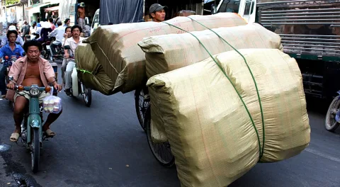 Ein Radfahrer zieht große, verschnürte Ballen durch eine belebte Straße, links fährt ein Motorrollerfahrer mit einem Paket auf dem Lenker.
