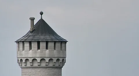 Oberer Teil eines runden Steinturms mit spitzem, dunklem Schieferdach, Zierkugel auf der Spitze und einer Bogengalerie unter dem Dach. Links der Turm, rechts eine leere graue Himmelsfläche.